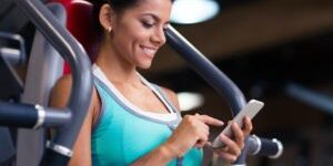 Portrait-of-a-cheerful-sports-woman-using-smartphone-in-fitness-gym